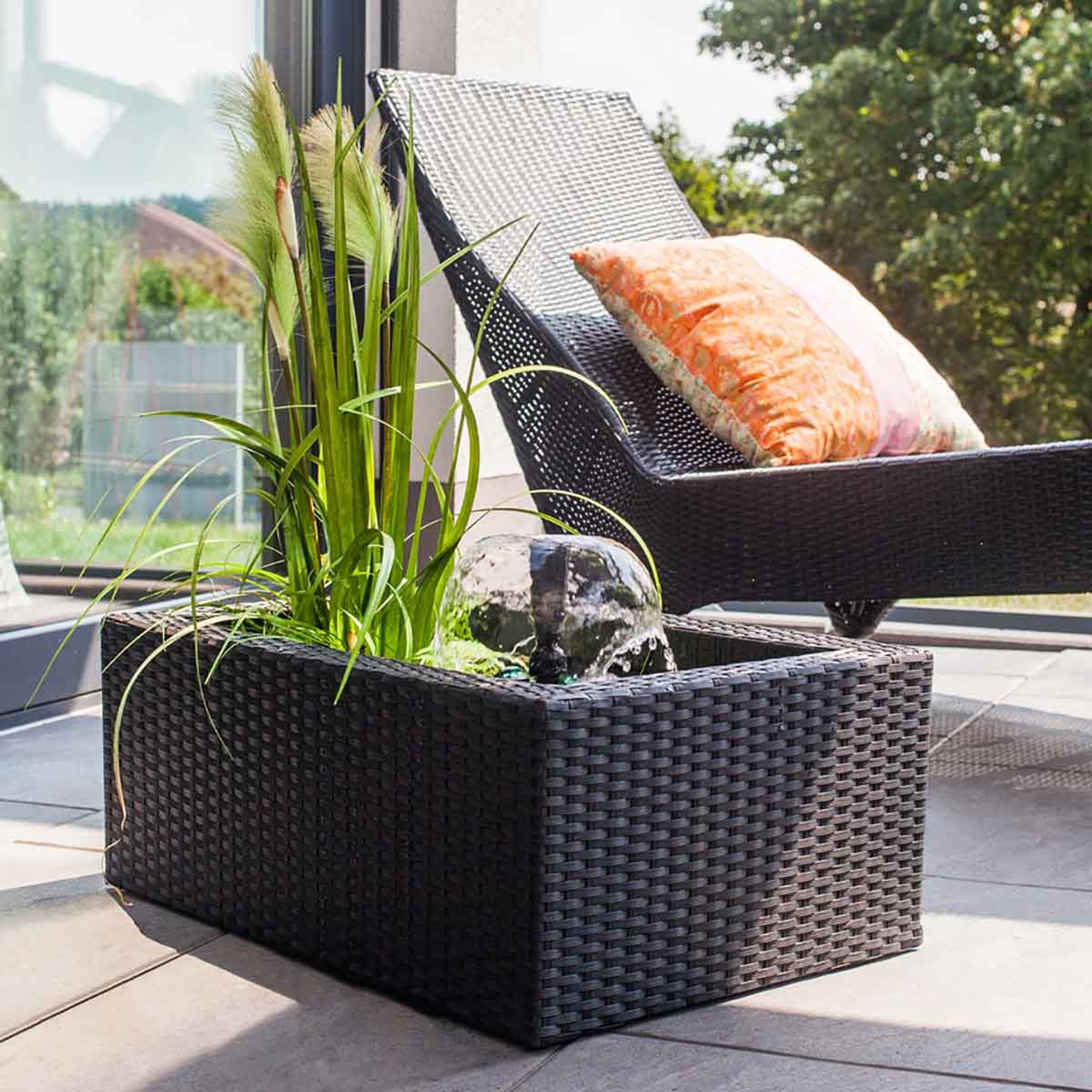 Heissner 015196-00 Rattan Terrassen-Hochteich mit Wasserspiel, schwarz bepflanzter grauer Gartenbrunnen aus Rattan neben Sonnenliege
